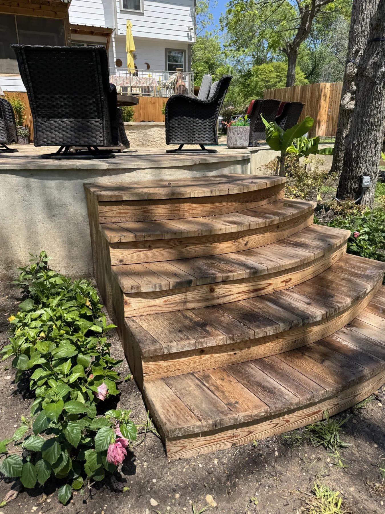 Curved wooden steps built up to a raised backyard patio, edged with plants